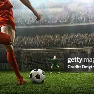 A male soccer player kicks a ball and soccer goalie jumping in motion for a ball while defending his gates on wide angle panoramic image of a outdoor soccer stadium or arena full of spectators under a sunny sky. The image has depth of field with the focus on the foreground part of the pitch. With intentional lensflares. Players are wearing unbranded soccer uniform.