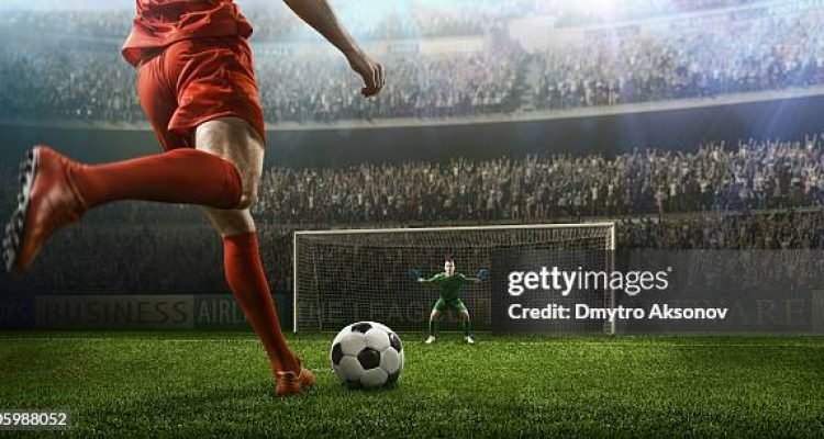A male soccer player kicks a ball and soccer goalie jumping in motion for a ball while defending his gates on wide angle panoramic image of a outdoor soccer stadium or arena full of spectators under a sunny sky. The image has depth of field with the focus on the foreground part of the pitch. With intentional lensflares. Players are wearing unbranded soccer uniform.